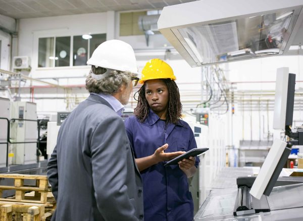 Serious black female factory worker talking to boss at machine on plant floor, pointing at tablet screen. Medium shot, copy space. Production process or machinery concept